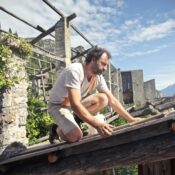 Side view of male builder in summer wear reconstructing rooftop and sitting on wooden beams of aged building on background of scenic landscape in sunny day