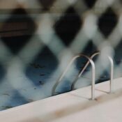 A view of a neglected swimming pool with a metal ladder, seen through a chain-link fence.