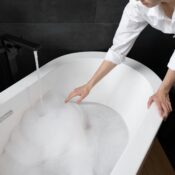 A woman in a bathroom runs water into a foam-filled bathtub, creating a relaxing atmosphere with a candle nearby.