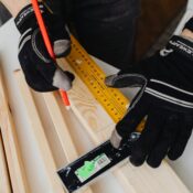 Close-up of a carpenter wearing gloves while measuring a wood plank with a ruler and pencil.