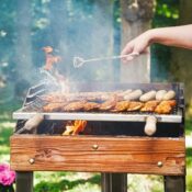 A person grilling meat at a backyard barbecue on a sunny day, with smoke rising and vibrant flowers nearby.