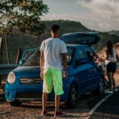 A young couple with a blue car on a scenic road, set against a beautiful mountain backdrop.
