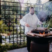 A chef skillfully grills meat on an open flame in a beautiful garden setting, surrounded by greenery.
