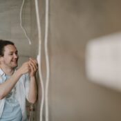 An electrician with curly hair adjusts an outlet indoors, smiling as he works.