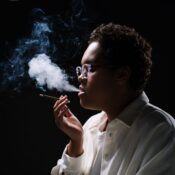Close-up of a woman in glasses exhaling smoke against a dark background.