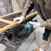 A carpenter using a power drill to work on wooden planks in an indoor workshop setting.