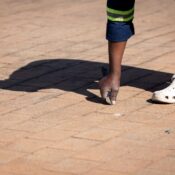 A worker in uniform repairs an outdoor brick pathway using a tool, wearing white clogs.