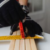 Close-up of a gloved carpenter measuring wood indoors using a ruler and pencil.