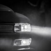 Black and white photo of a car headlight illuminating through fog at night.