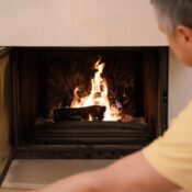 A man in a yellow shirt tends to the burning fire in a cozy indoor fireplace.