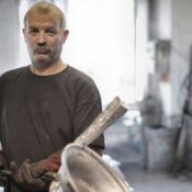 Serious middle aged turner in black t shirt and brown heat resistant gloves working with metal in light workshop and looking at camera