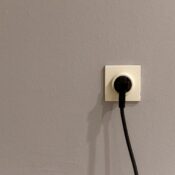 A black plug inserted into a white wall socket on a plain grey wall.